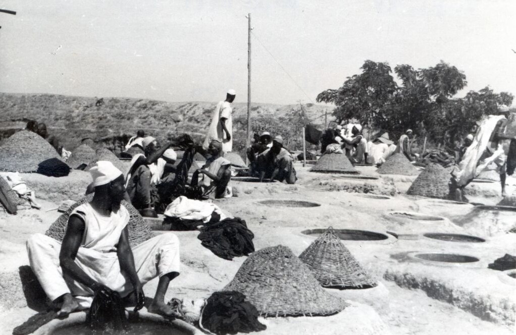 A field of circular dye pits cut into bare ground at Kano. Men in white caps crouch at the rims with dark wet cloth; conical woven covers stand between the pits, and dyed lengths hang drying in the background.