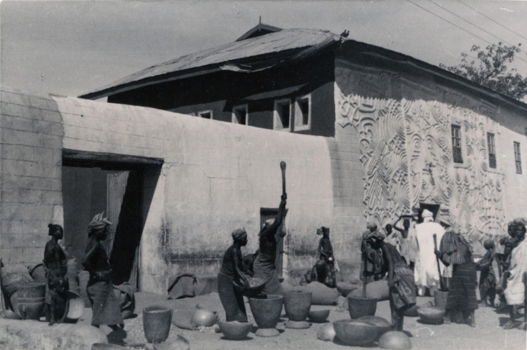 Women outside a compound wall grinding groundnuts with long pestles in wide mortars, surrounded by large pots and bowls. A decorated building stands behind them.